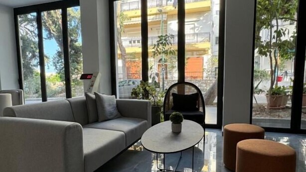 Un salon moderne avec un canapé, une table basse et deux poufs. De grandes fenêtres offrent une vue sur l’extérieur.