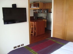Un dormitorio con cama, televisor montado en la pared, zona de cocina con armarios de madera y un refrigerador, y una pequeña mesa de comedor con sillas.