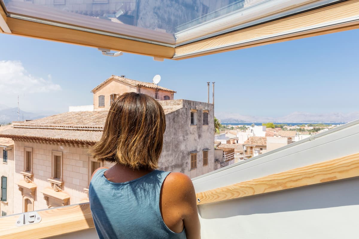 A person standing by a window overlooking a cityscape with buildings and mountains in the distance.