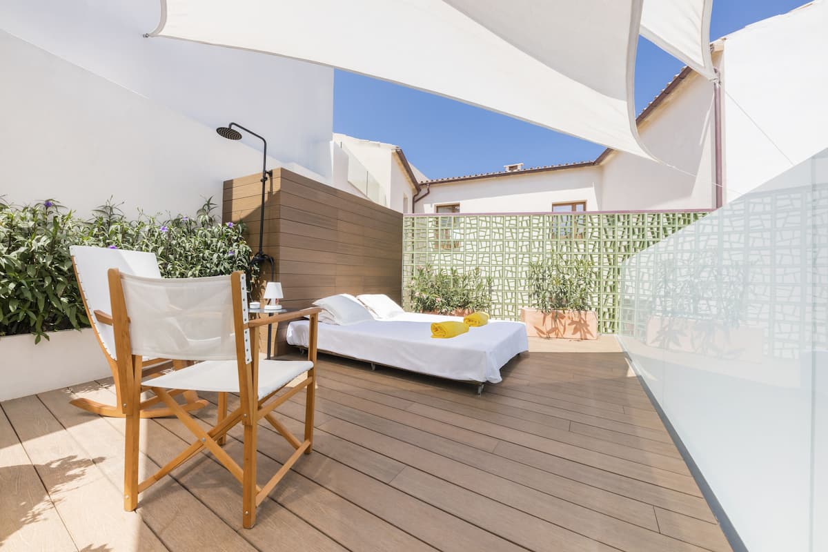 A sunlit outdoor balcony with a wooden deck, a white chair, a small table, and a bed with a white cover.