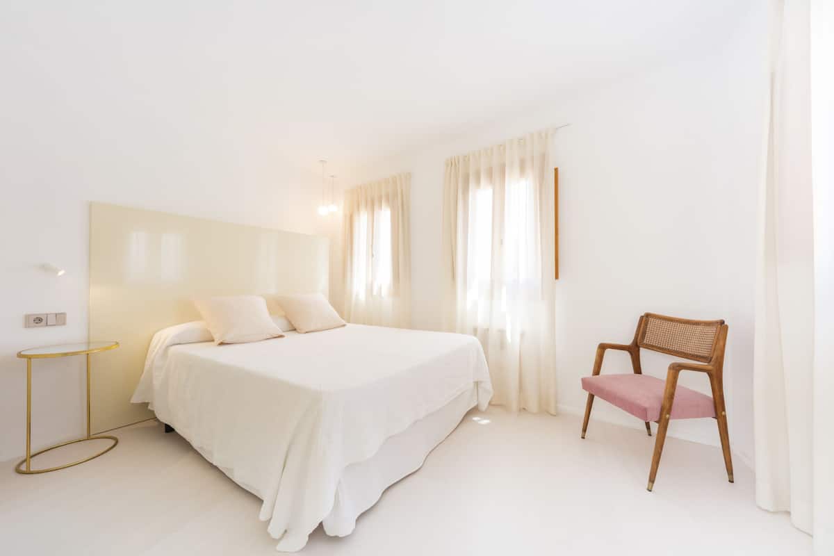 A minimalist bedroom with a large bed, a small round table, and a wooden chair.