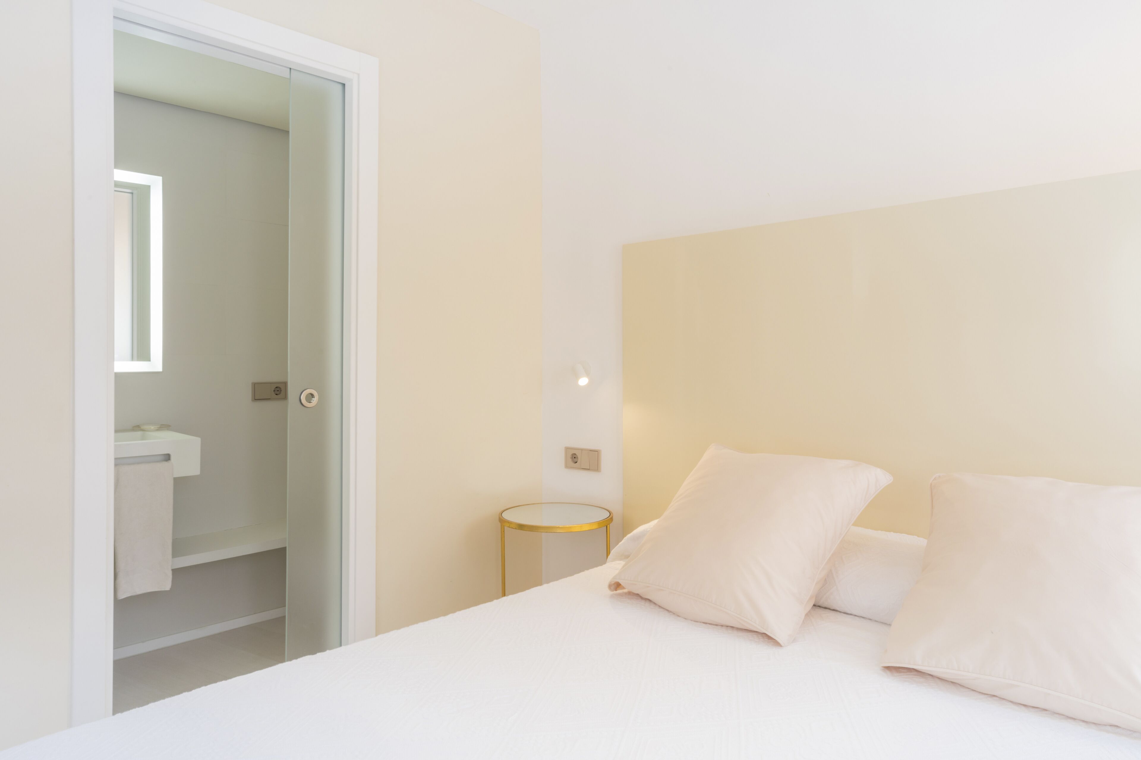 A modern hotel room with a bed, a round side table, a small round mirror, and a bathroom visible through an open door.