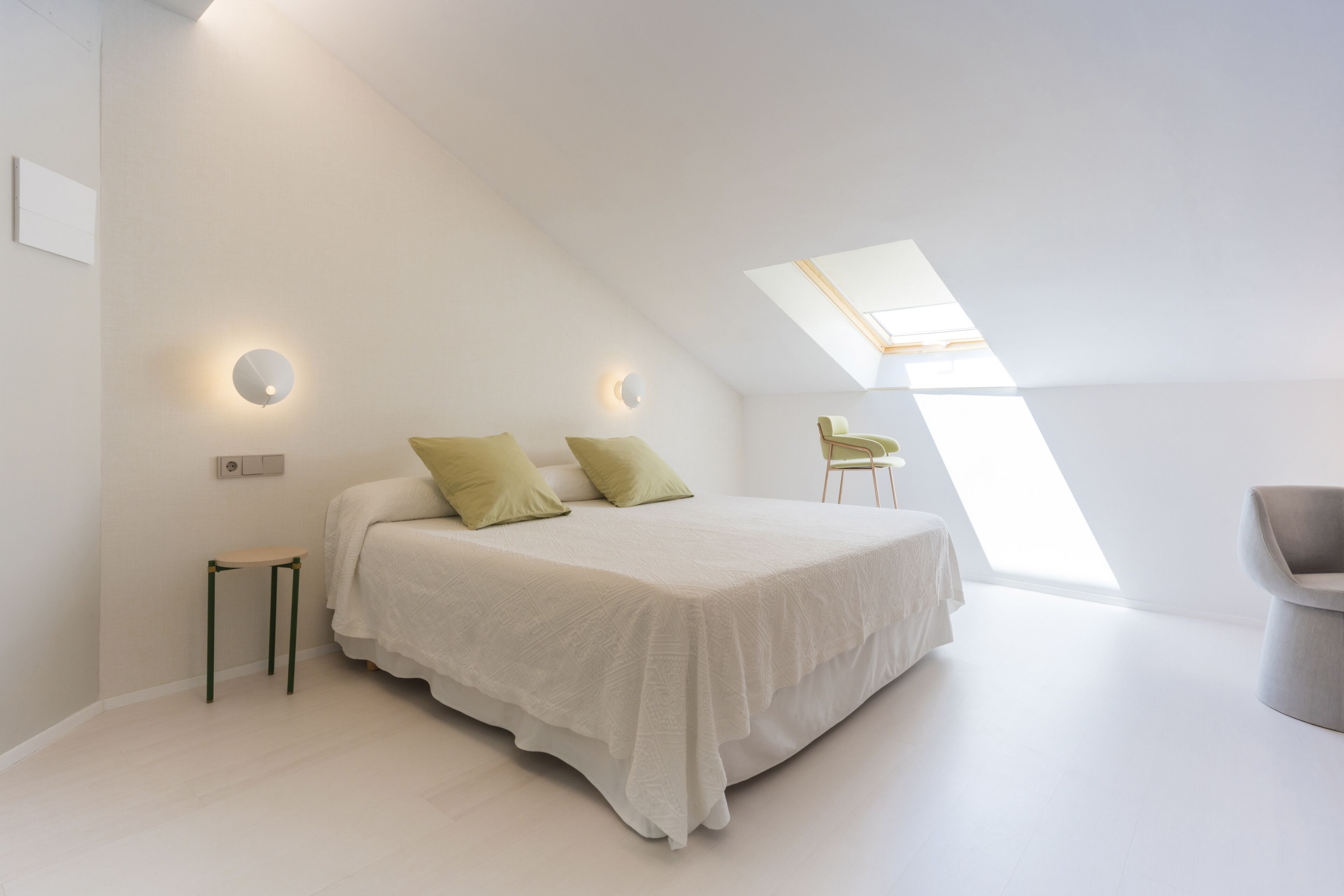 Un dormitorio moderno con una cama grande, una mesita pequeña, una silla y un tragaluz.