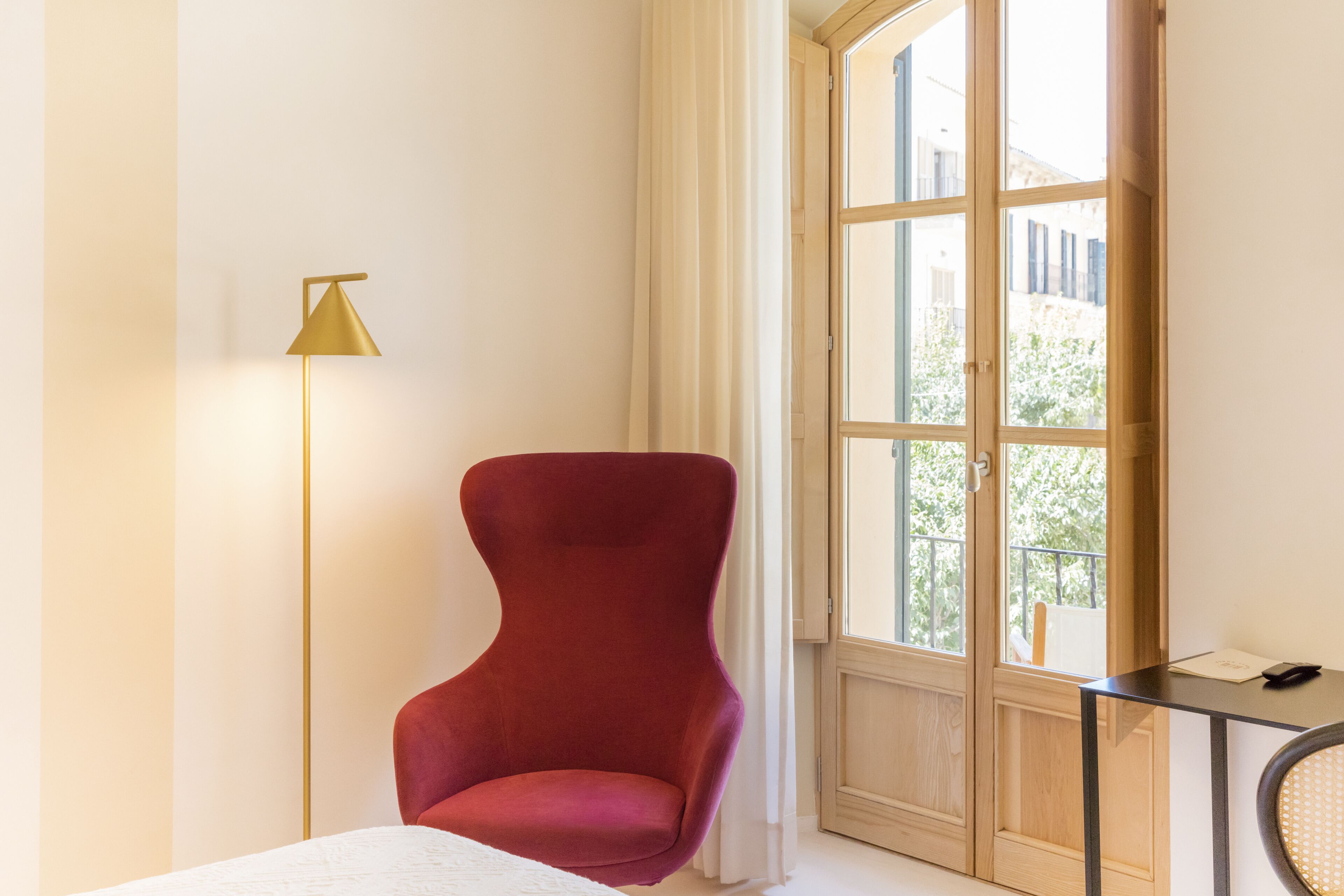 A room with a red chair, a floor lamp, a window with a view of buildings, and a small table with a phone and papers.
