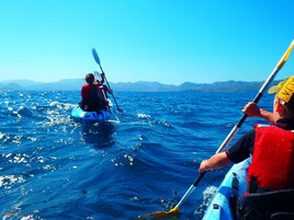 Dos kayakistas en un kayak azul sobre un mar tranquilo con montañas a lo lejos.