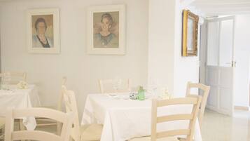Un espace repas avec des tables et des chaises blanches, des portraits encadrés accrochés au mur et une porte donnant sur une autre pièce.
