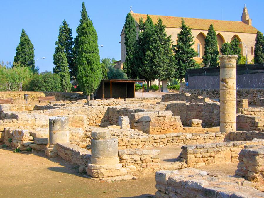 Ruinas antiguas con columnas y muros de piedra, un edificio con arcos al fondo y árboles alrededor del sitio.