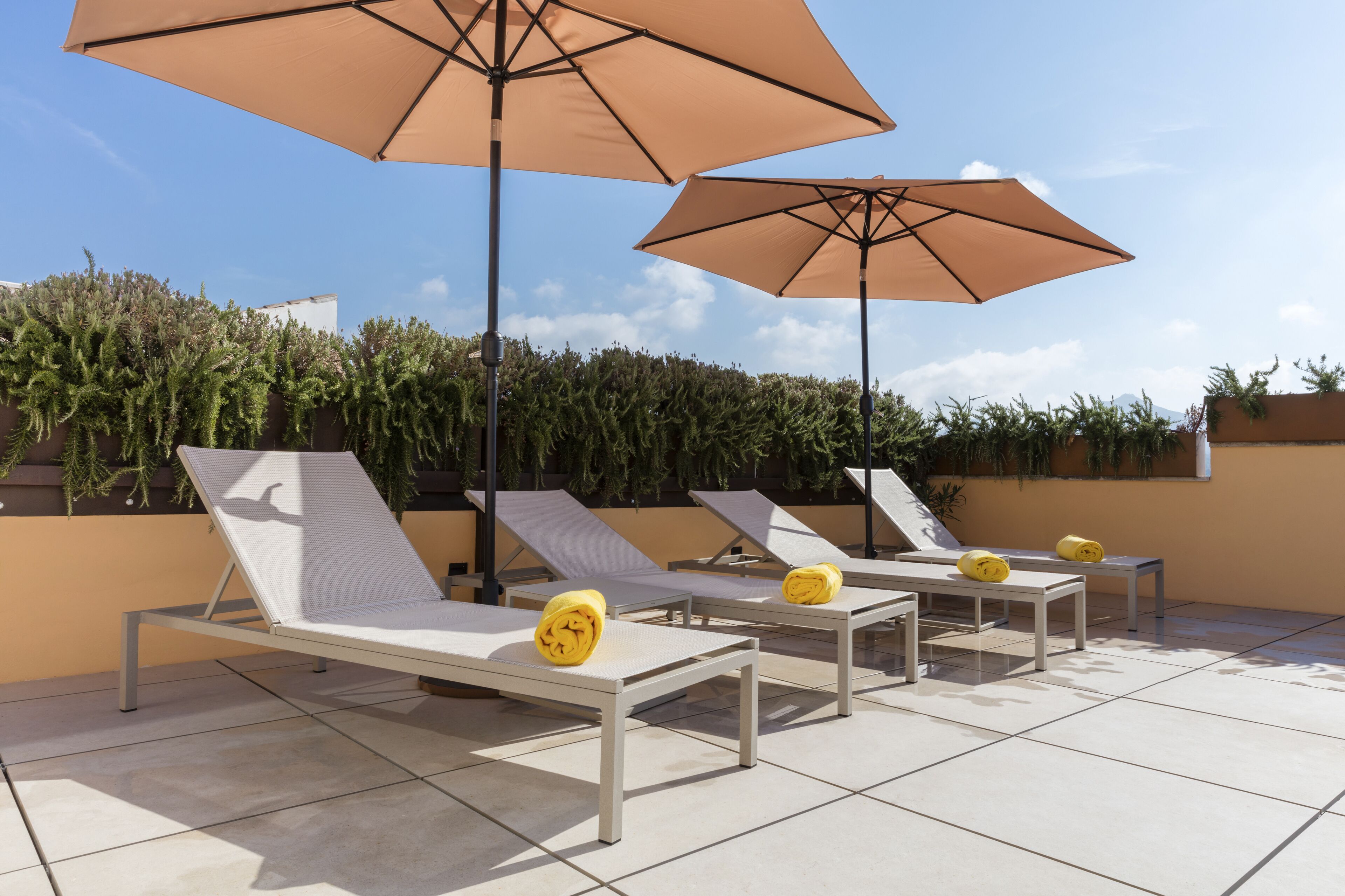 A rooftop lounge area with white lounge chairs and beige umbrellas, surrounded by greenery.