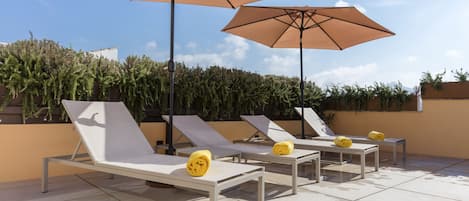 A rooftop lounge area with white lounge chairs and beige umbrellas, surrounded by greenery.