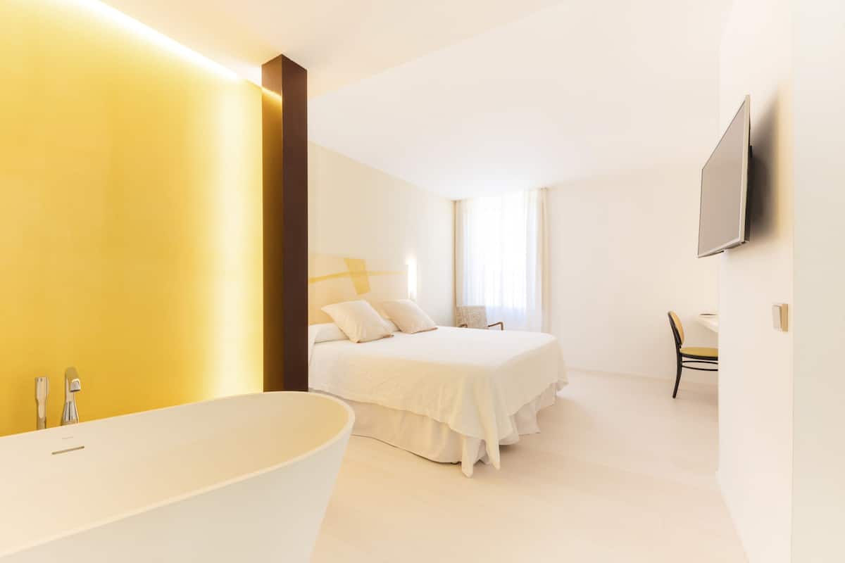 A modern bedroom with a large bed, a white bathtub, and a flat-screen TV mounted on the wall.