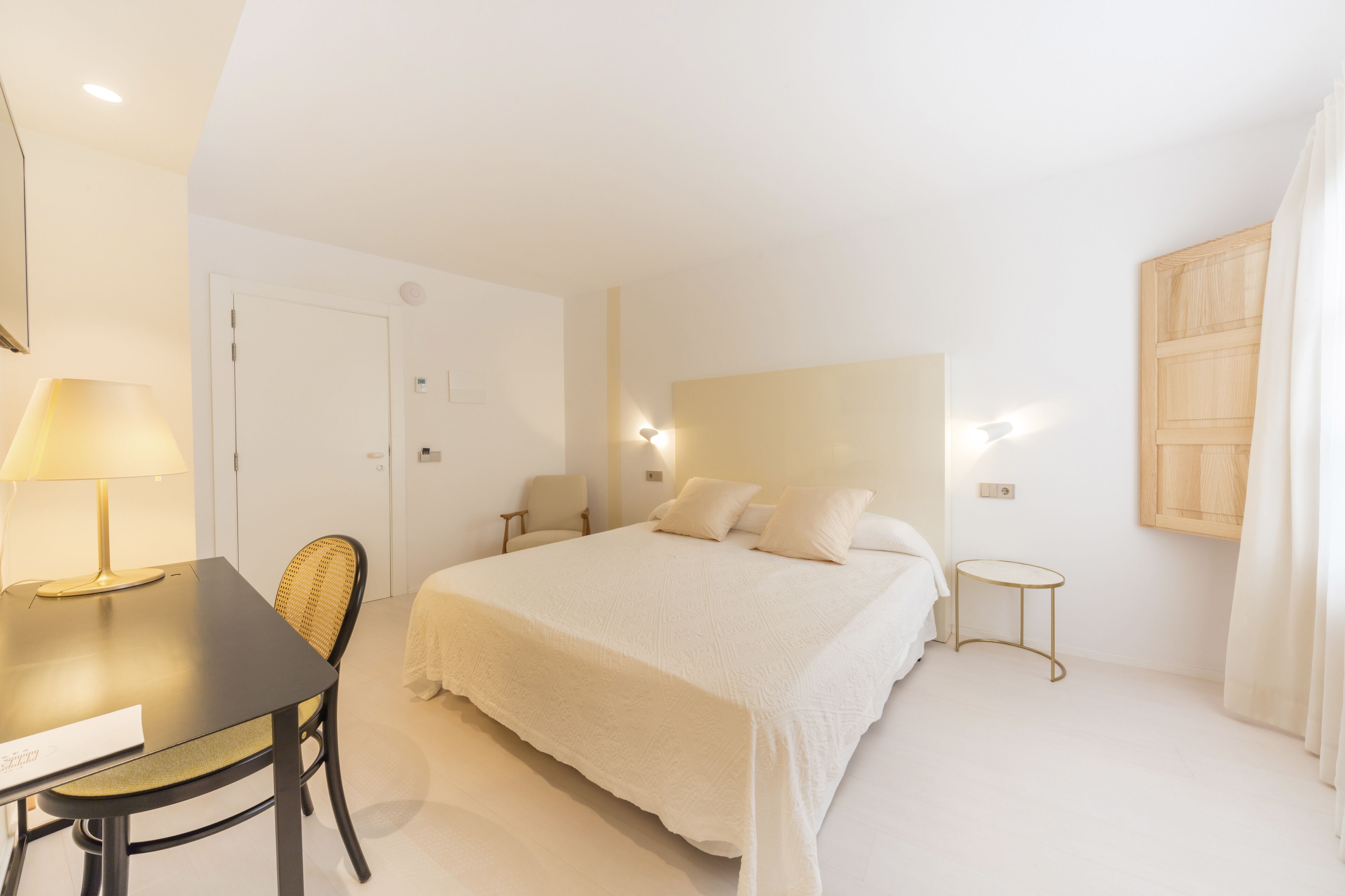 A hotel room with a bed, a desk, a chair, a small table, and a wall-mounted light fixture.