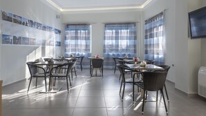 A modern dining area with multiple tables and chairs, large windows with blinds, and a wall-mounted television. - Hotel Sonia (Kos)