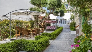 An outdoor patio with wicker furniture and a white umbrella, surrounded by neatly trimmed hedges and flowering plants. - Hotel Sonia (Kos)
