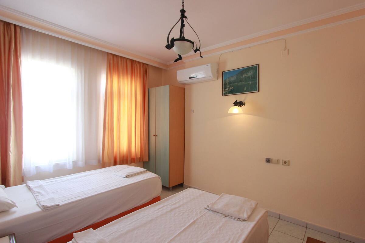 Habitación de hotel con dos camas individuales, un ventanal con cortinas, un equipo de aire acondicionado, una lámpara de pared y una puerta.