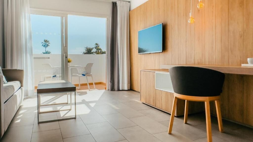 A modern hotel room with a balcony, a dining area, and a TV mounted on the wall.