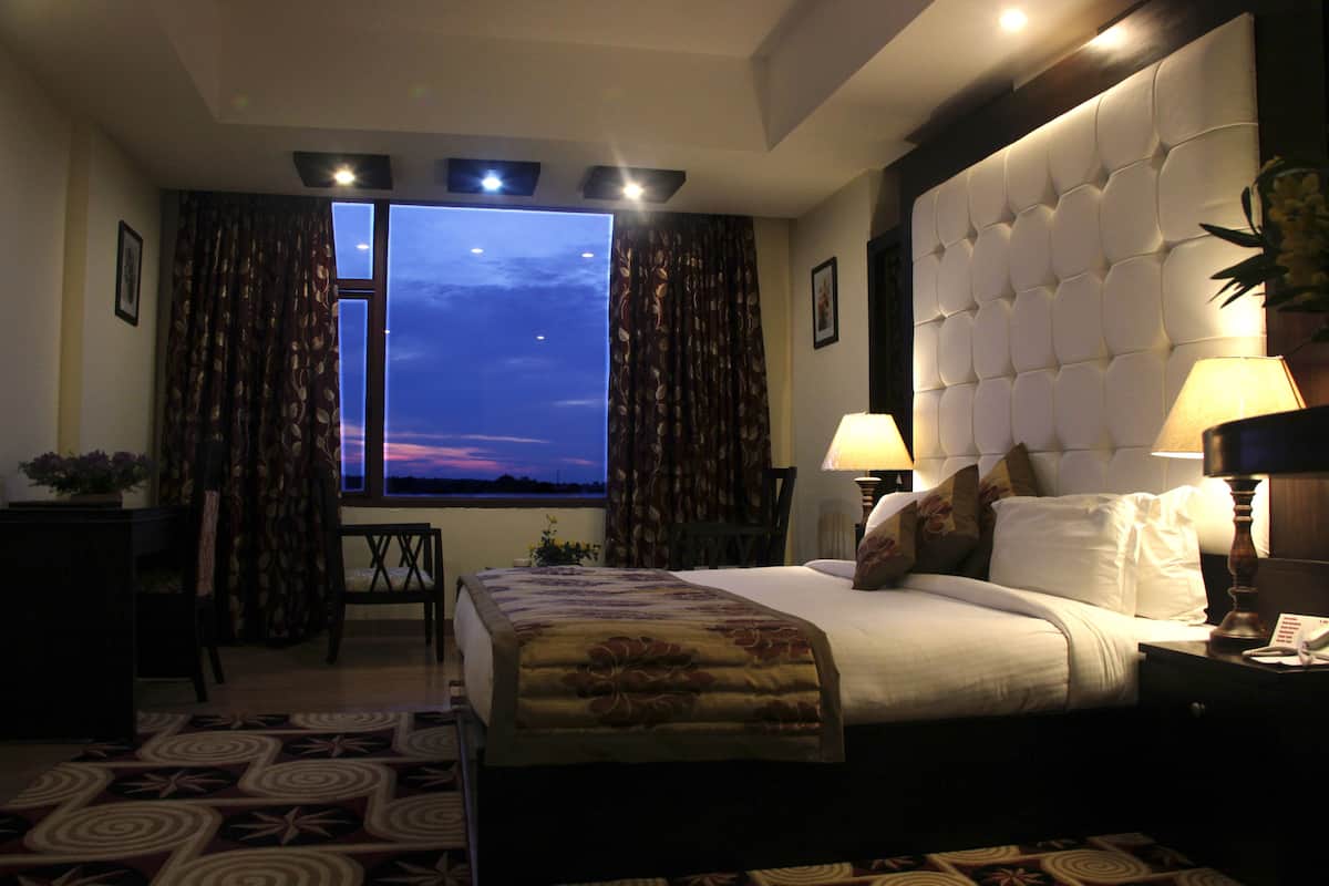 A hotel room with a large bed, a nightstand with a lamp, a desk with a chair, and a view of the sky through the window.
