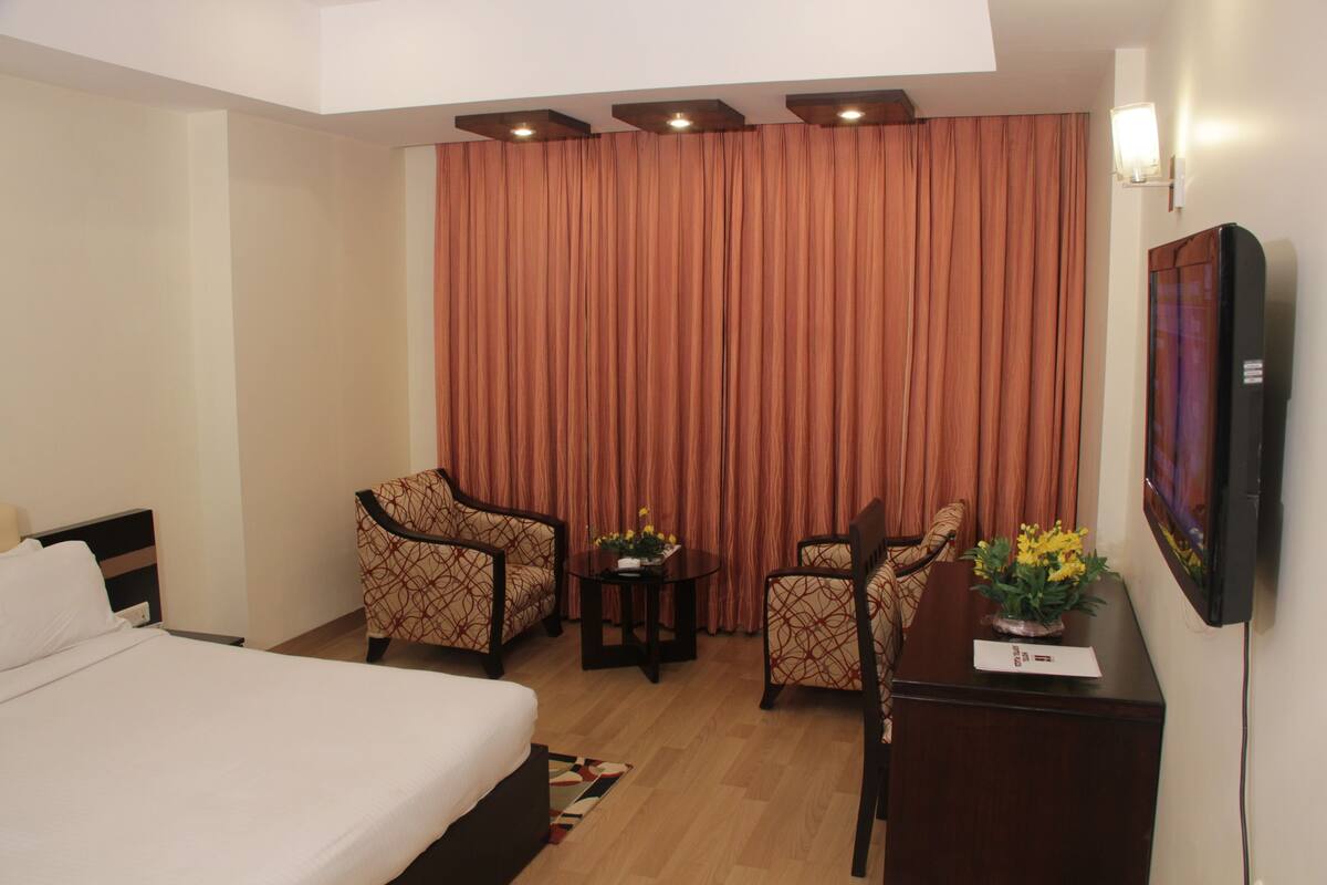 A hotel room with a bed, two armchairs, a small table with flowers, and a TV on the wall.