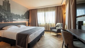 A hotel room with a bed, a desk, a chair, a TV, and a mural of a cityscape.