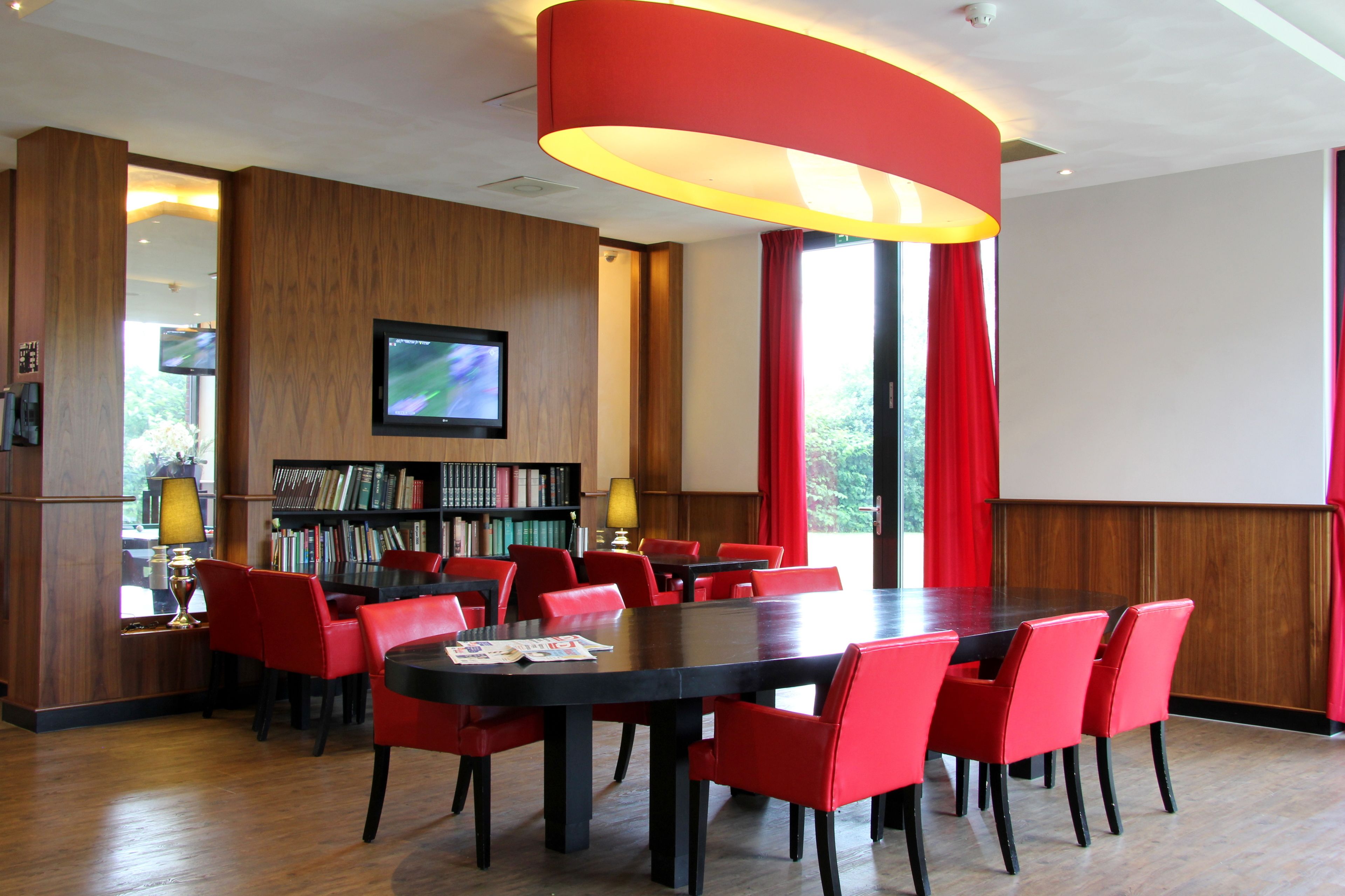 A conference room with a large table, red chairs, a bookshelf, and a television screen.