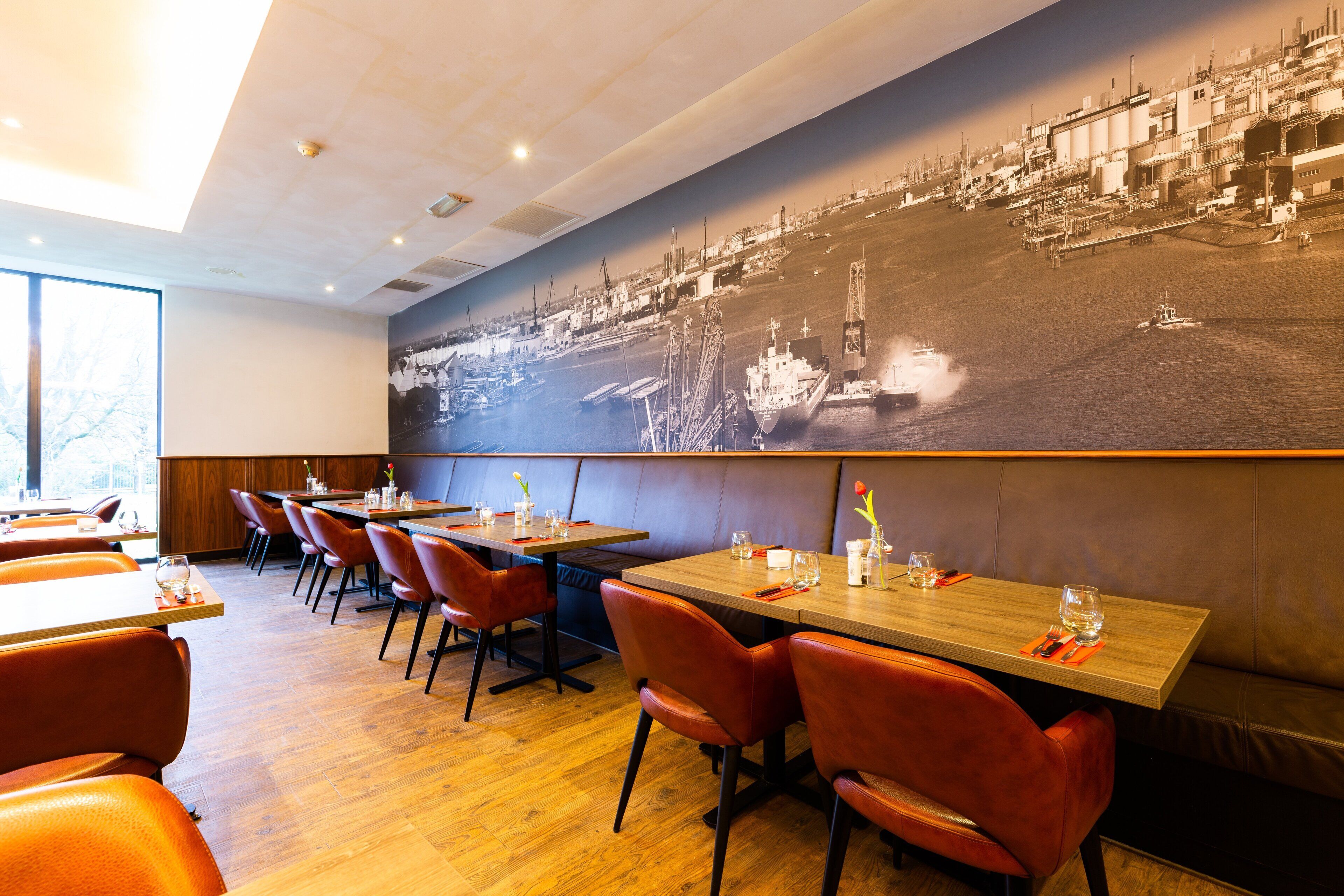 A restaurant with wooden tables and red chairs, a large black and white mural on the wall, and a view of the water outside.