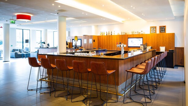 A modern bar area with a long counter, multiple stools, and a television screen.
