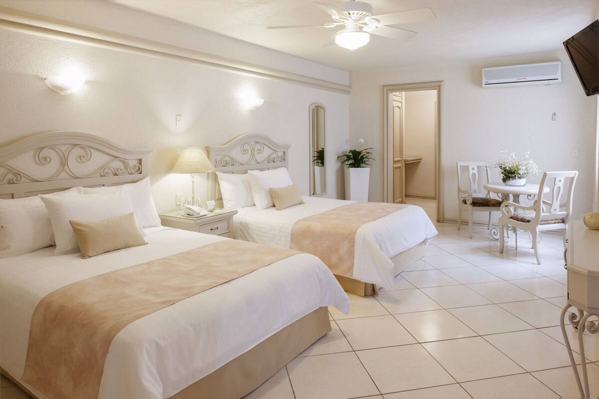 A hotel room with two beds, a desk, a chair, a mirror, and a ceiling fan.