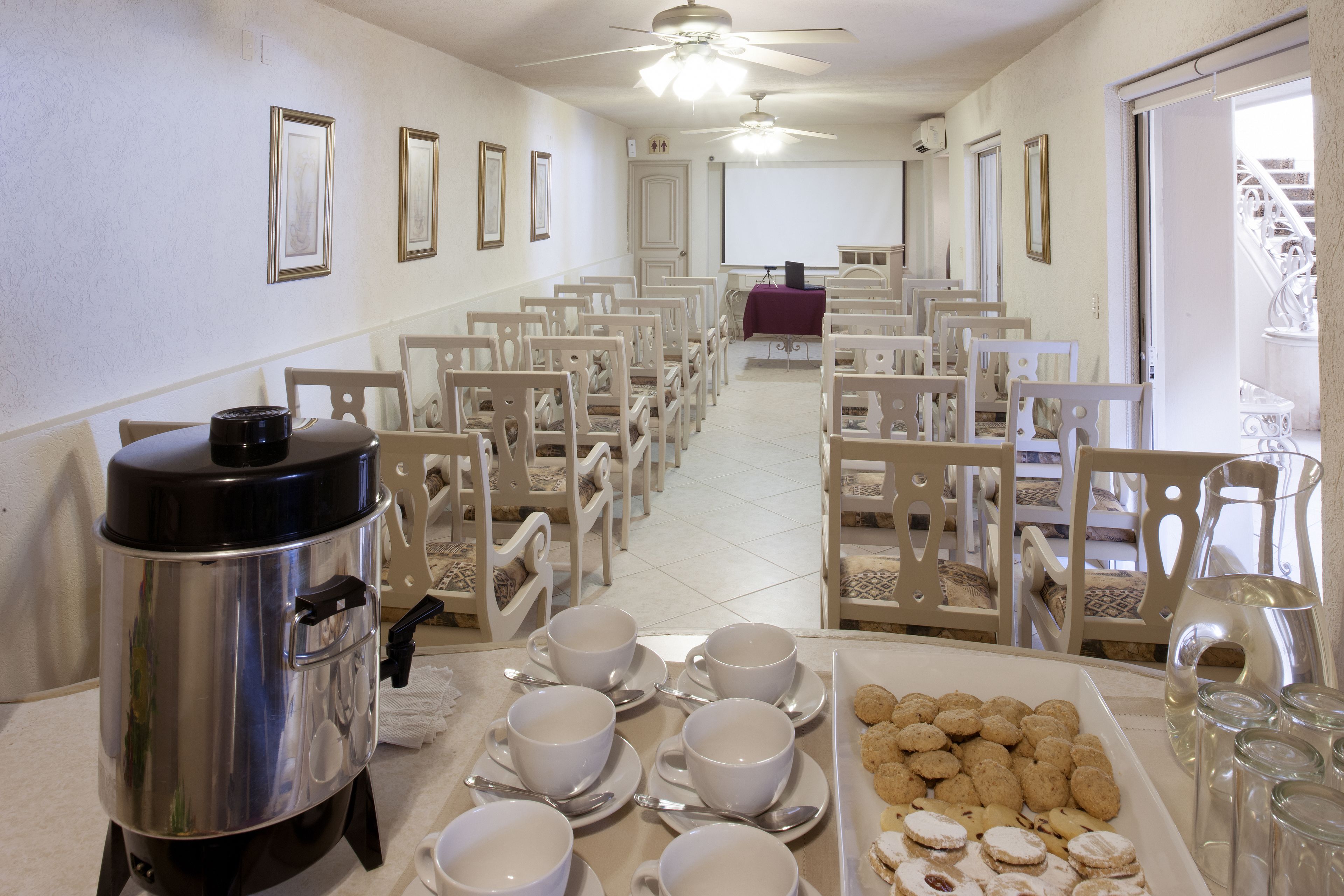 A hotel dining area with rows of white chairs, a buffet table with white cups and saucers, a silver food warmer, and a tray of cookies.
