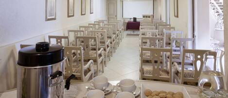 A hotel dining area with rows of white chairs, a buffet table with white cups and saucers, a silver food warmer, and a tray of cookies.