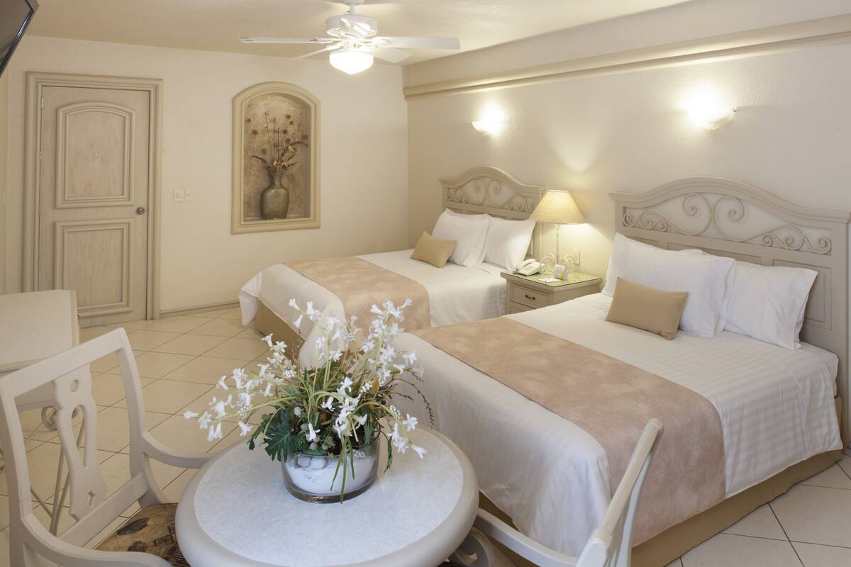 A hotel room with two beds, a round table with a vase of flowers, a ceiling fan, and a painting on the wall.