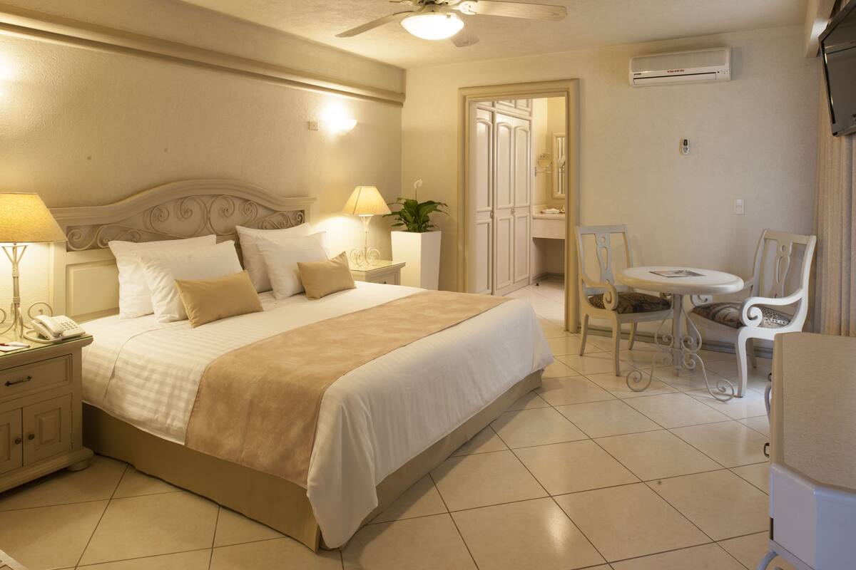 A hotel room with a large bed, bedside lamps, a small table with chairs, and an air conditioning unit on the wall.