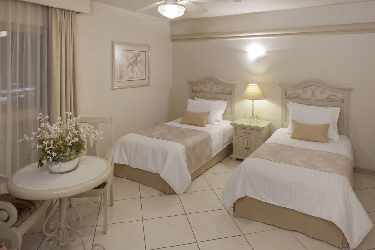A hotel room with two beds, a small table with a vase of flowers, a chair, and a bedside lamp.