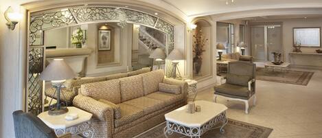 A spacious living area with a sofa, armchairs, and a coffee table, featuring a decorative mirror, a ceiling fan, and a staircase in the background.
