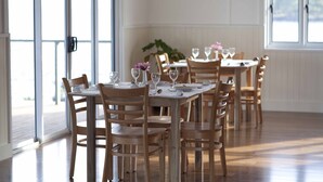 A dining area with wooden tables and chairs, large windows, and a plant in the background. - Madang Lodge Hotel (Madang)