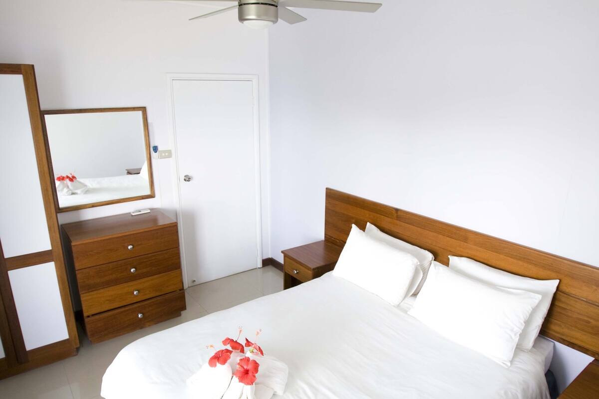A neatly made bed with white linens and a floral arrangement, a wooden nightstand, a dresser, a ceiling fan, and a white door.
