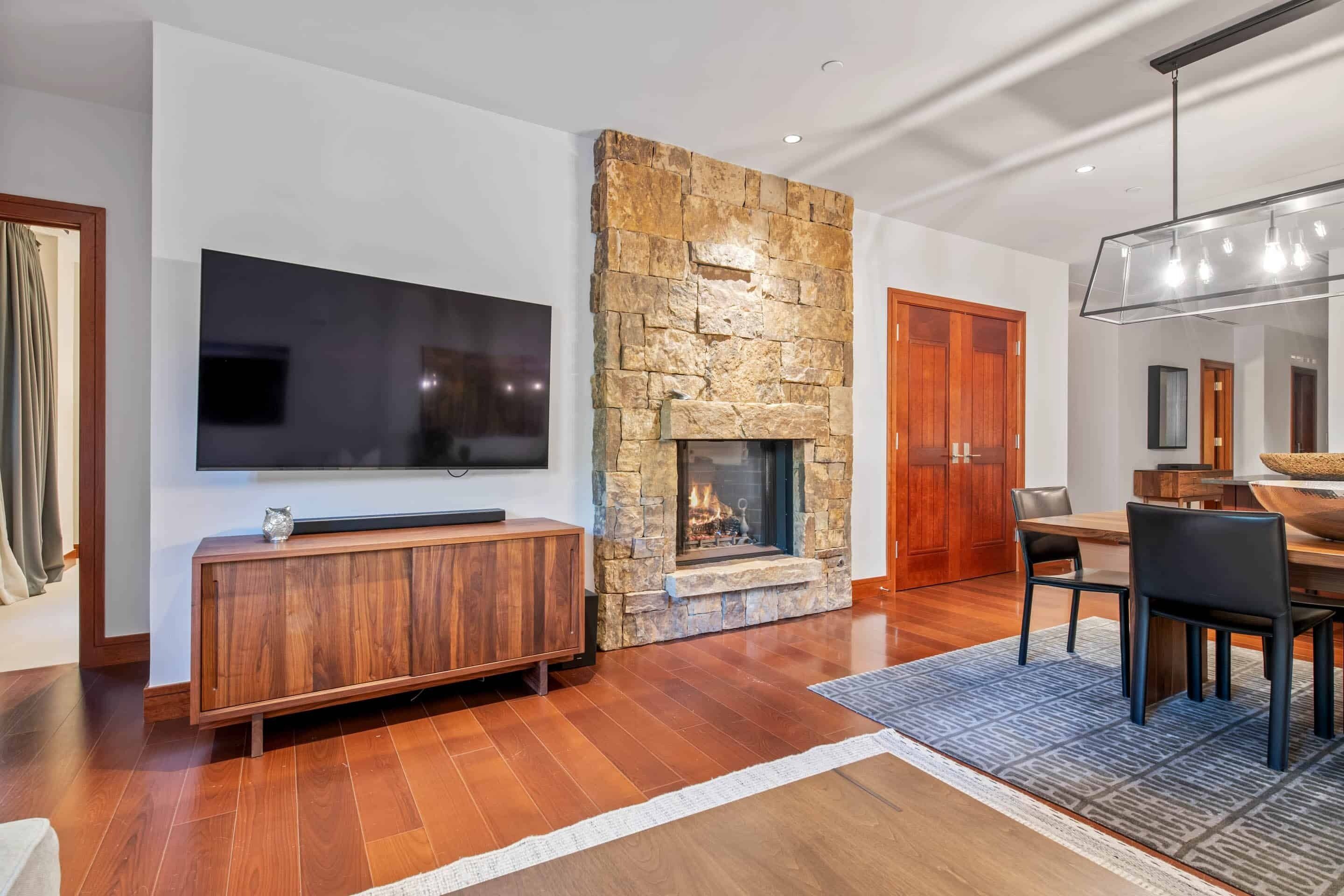 Una sala de estar moderna con chimenea de piedra, televisor de pantalla plana montado en la pared, mueble de entretenimiento de madera y zona de comedor con una mesa grande y sillas.