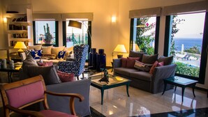 A modern living room with a sofa, armchairs, a coffee table, and a bookshelf. Large windows offer a view of the ocean. - Kalidon Panorama (Samos)