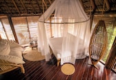 A room with a bed under a canopy, a wooden floor, a wicker chair, and a round table with a vase.