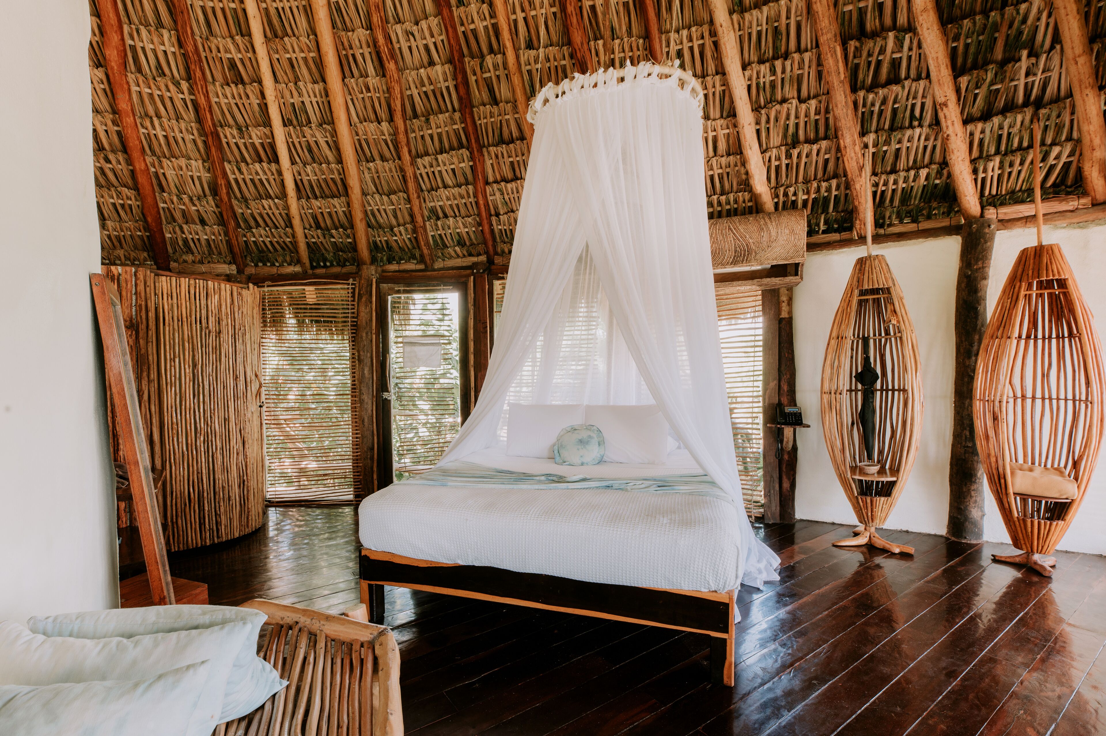 A room with a bed under a thatched roof, wooden furniture, and woven chairs.