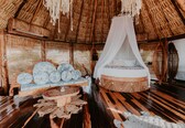 A thatched-roof room with a bed, a wooden bench, and woven mats.