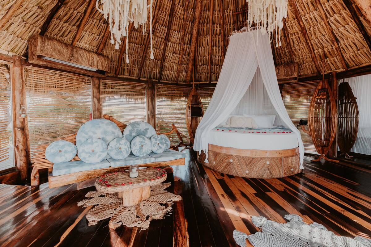 A thatched-roof room with a bed, a wooden bench, and woven mats.