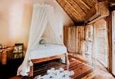 A room with a wooden bed, a canopy, a wooden floor, and a thatched roof.