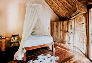 A room with a wooden bed, a canopy, a wooden floor, and a thatched roof.