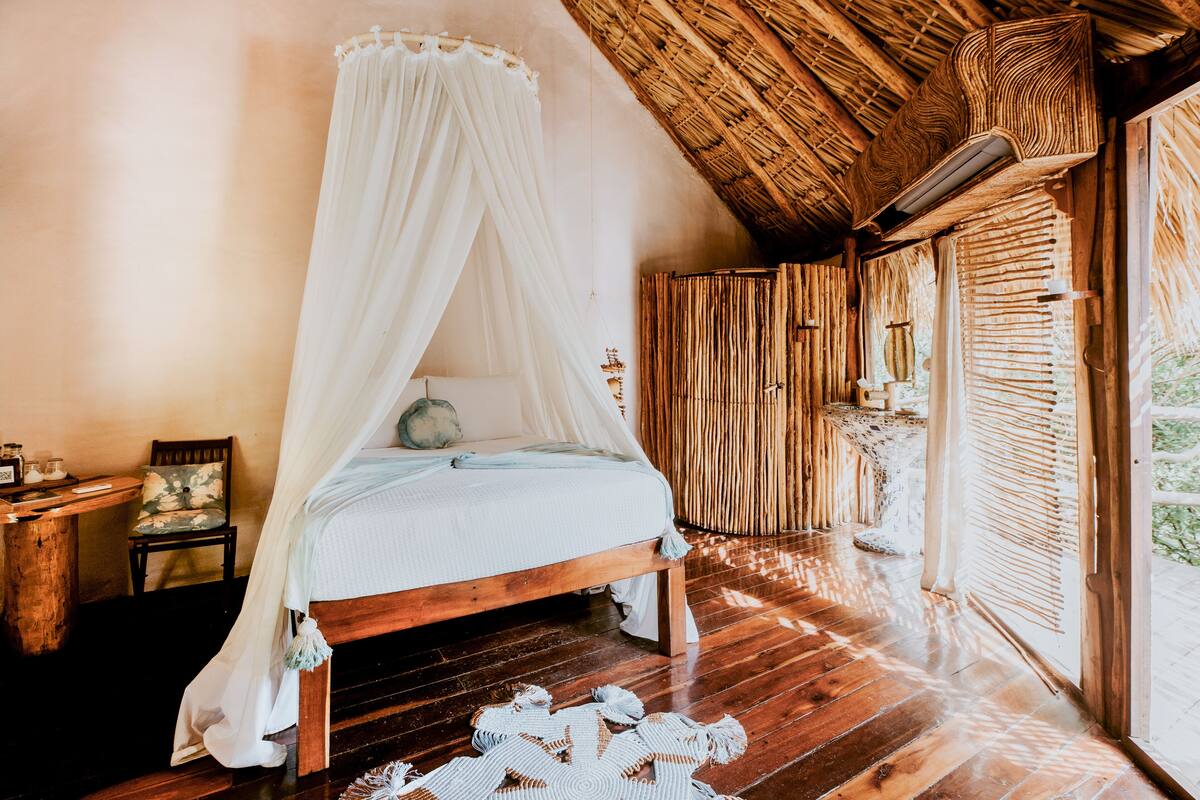 A room with a wooden bed, a canopy, a wooden floor, and a thatched roof.