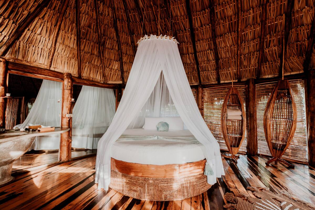 A circular wooden bed with a white canopy, wooden floors, and a thatched roof.