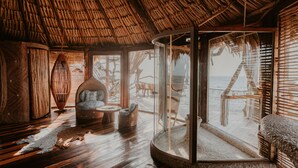 Una habitación con techo de paja, piso de madera, una zona acristalada con vista a la playa y una silla de mimbre.