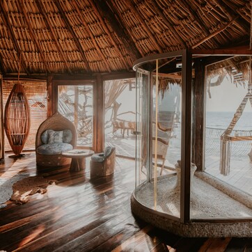 A room with a thatched roof, wooden floors, a glass enclosure with a view of the beach, and a woven chair.