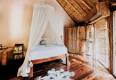 A room with a wooden bed, a canopy, a wooden floor, and a thatched roof.