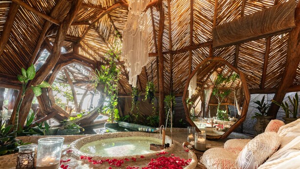 Una habitación con techo de paja, un jacuzzi rodeado de plantas y una cama con sábanas blancas.