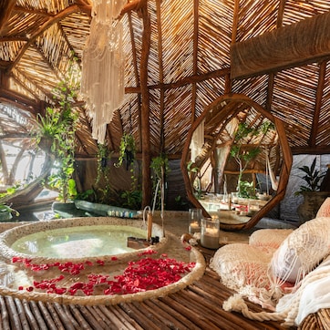 A room with a thatched roof, a hot tub surrounded by plants, and a bed with white linens.