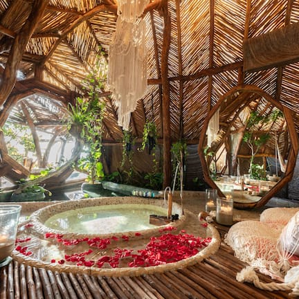 A room with a thatched roof, a hot tub surrounded by plants, and a bed with white linens.
